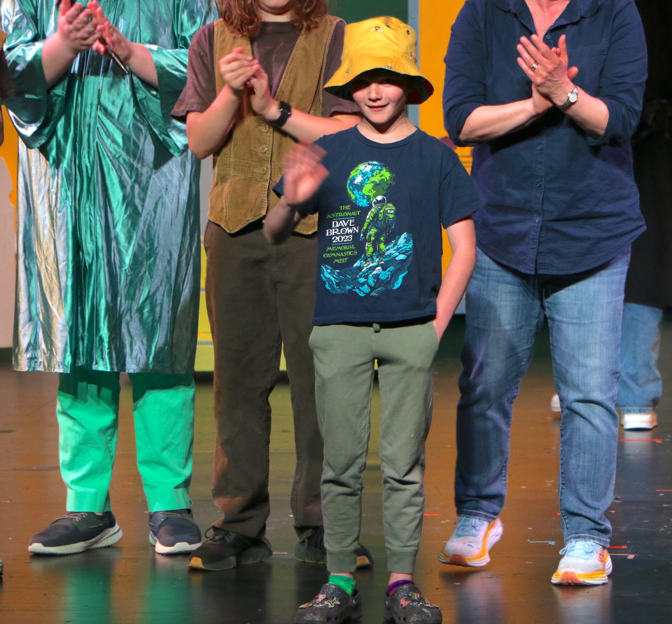 Students performing on stage - one in the middle with a yellow hat with others clapping around them